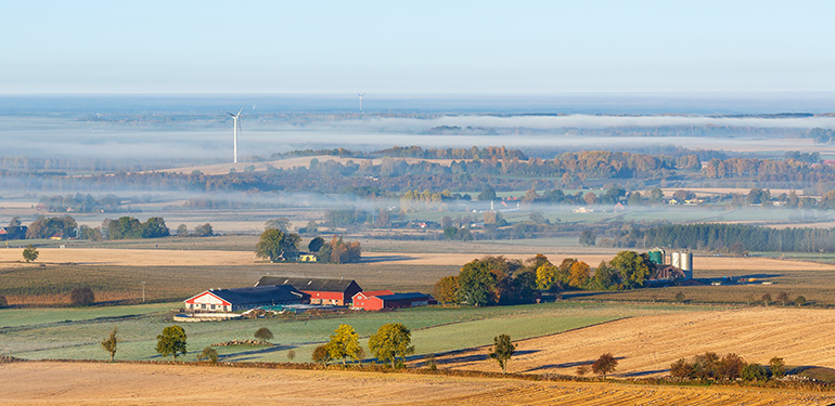 Drönarbild över ett svenskt lantligt landskap med fält, en gård och vindkraftverk