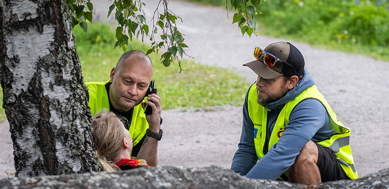 Två civila insatspersoner hjälper skadad kvinna som sitter lutad mot ett träd
