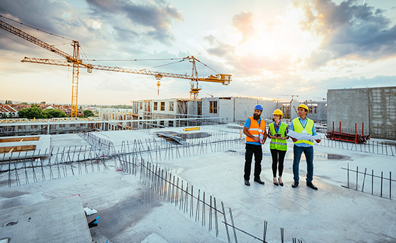 Byggarbetsplats, tre personer i skyddskläder diskuterar med varandra utifrån en ritning som en av dem håller i