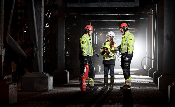 Tre personer i arbetskläder står i en dunkel gruva och diskuterar. Den ena personen har en handbrandsläckare i handen.