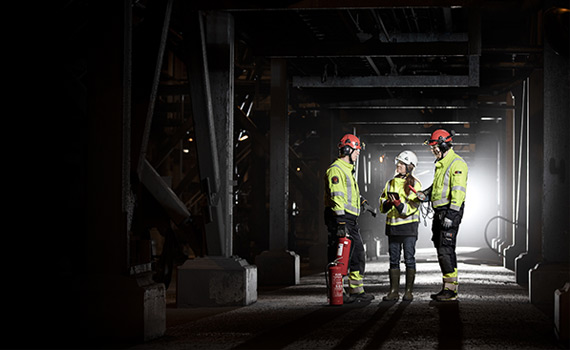 Tre personer i arbetskläder står i en mörk gruva och diskuterar något. En av personerna håller i en brandsläckare.