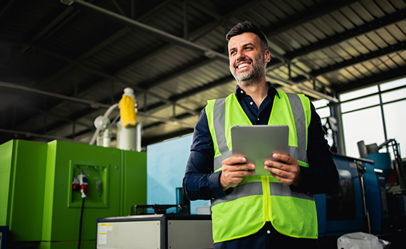 Man med skjorta och gul väst står i industriell lokal med surfplatta och ler