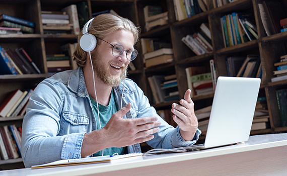 En man sitter med hörlurar i ett bibliotek med sin laptop och pratar med någon i ett videosamtal