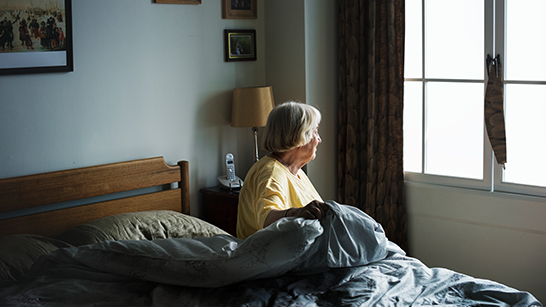 Äldre kvinna sitter i en säng och ser ut genom fönstret