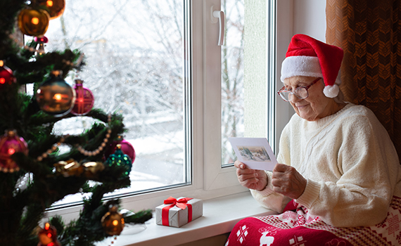 Äldre kvinna sitter intill en julgran och fönster och läser på ett kort