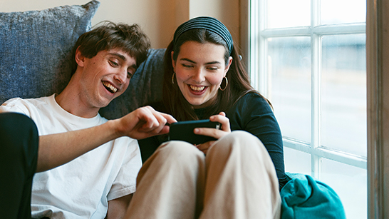 Två ungdomar sitter bredvid varandra och skrattar medan de håller i en mobiltelefon