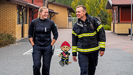 Två personer från räddningstjänsten går över en skolgård med figuren Flammy